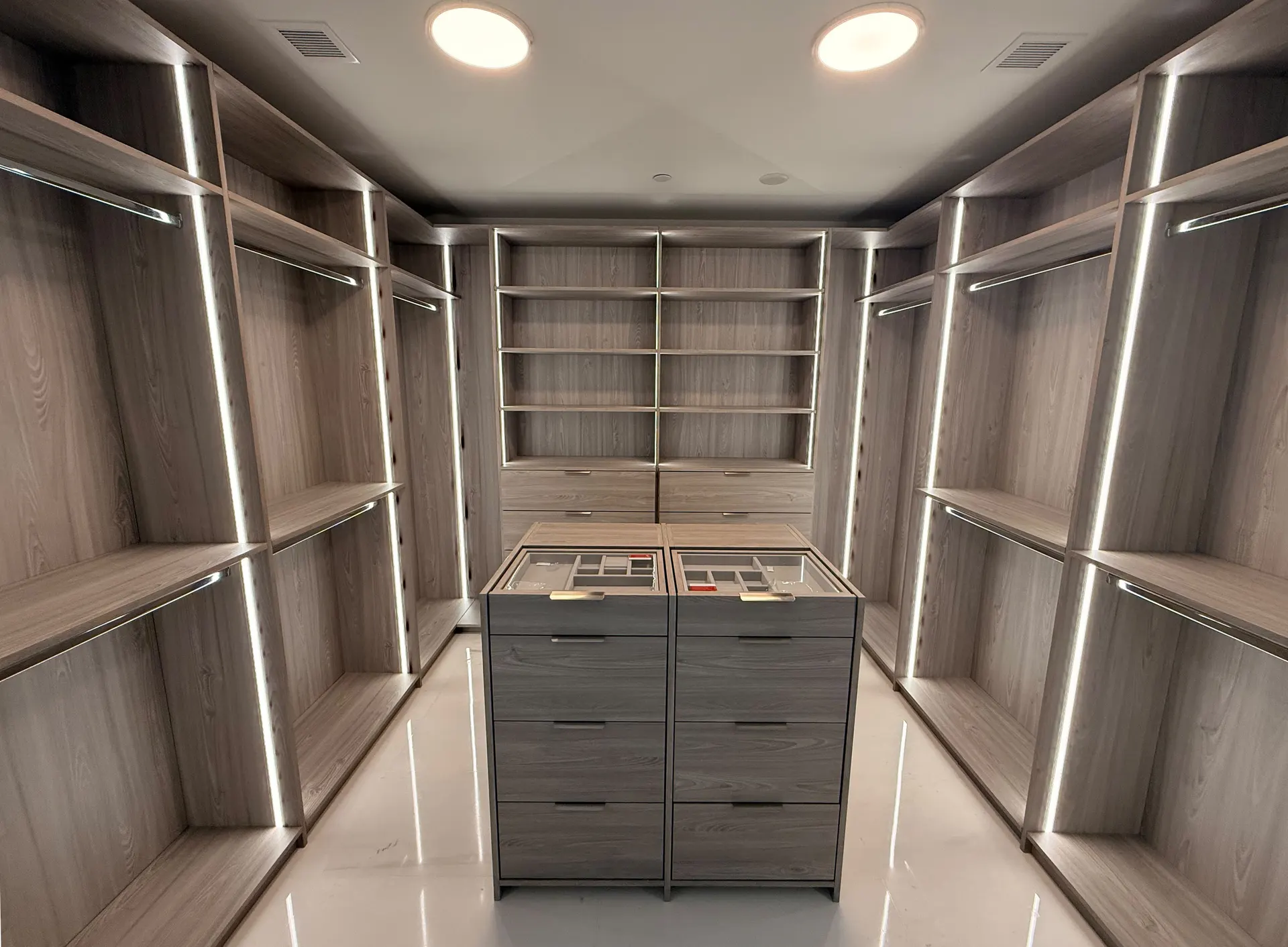 Walk-in closet in grey oak with lit hanging rails down both walls and a central island of drawers with glass-topped jewellery trays