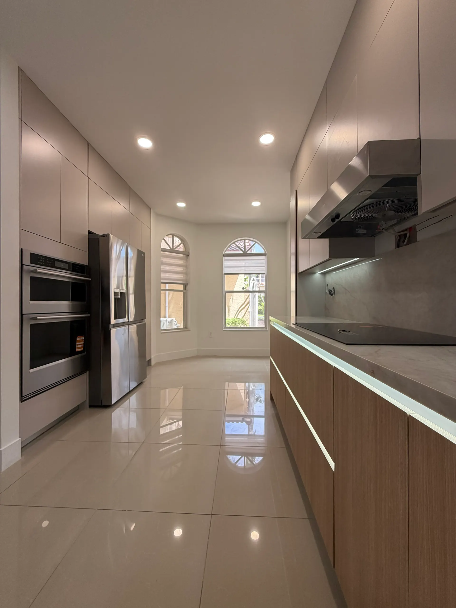 Galley kitchen with a run of walnut base cabinets under a lit marble counter, a stainless hood over the cooktop, and a wall of handleless cabinets around a built-in double oven and fridge