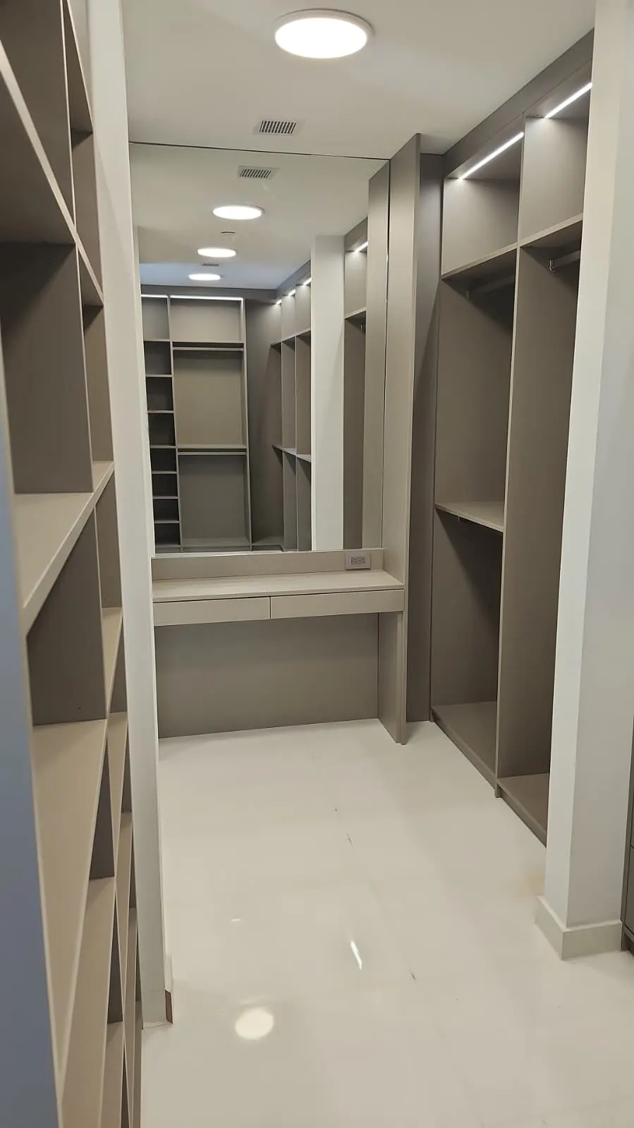 Walk-in closet in greige with open shelving and hanging rails on both sides, LED strips overhead and a dressing table with a wide mirror at the end