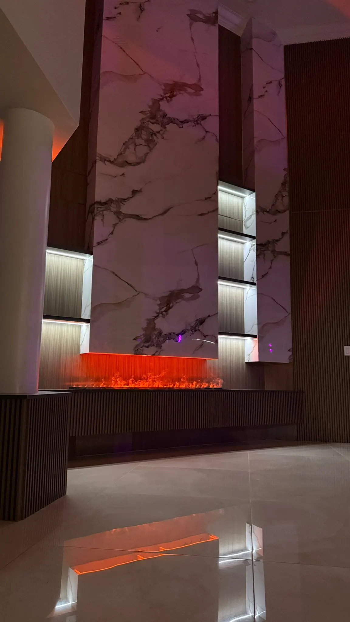Remodeled feature wall in marble-look panels and fluted wood, with lit niches and an electric fireplace glowing below